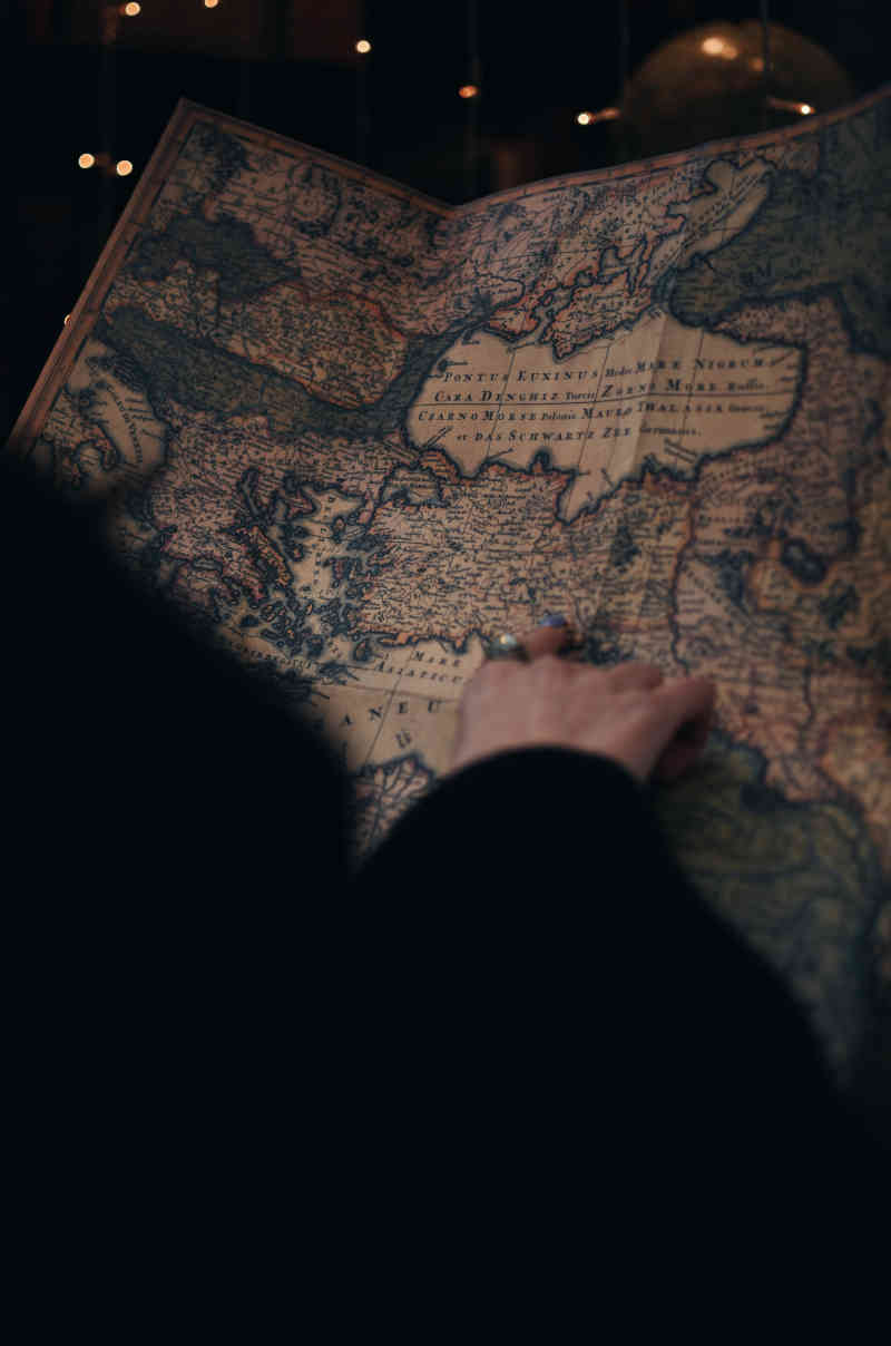Somebody in darkness whose visible hand rests on an antique parchment map, pointing with a ringed finger to a location on it.